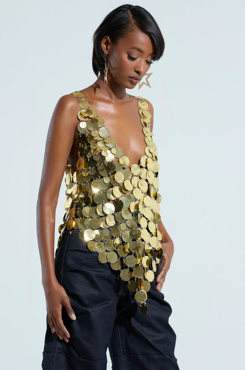 Sporting the GET ME BODIED GOLD SEQUIN TOP with its sleeveless design and V-neckline, a person pairs it flawlessly with dark pants for an elegant night-out ensemble. With short, dark hair and star-shaped earrings, they stand against a light background.