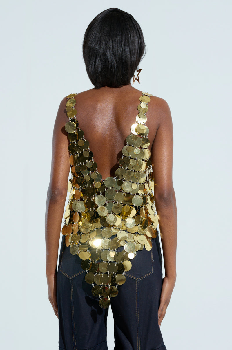 A person with short black hair is wearing the GET ME BODIED GOLD SEQUIN TOP, which features a sleeveless design and a v-neckline, perfect for a shimmering night-out look. Facing away to showcase its stunning design, they effortlessly pair it with dark pants that have visible stitching for added flair.