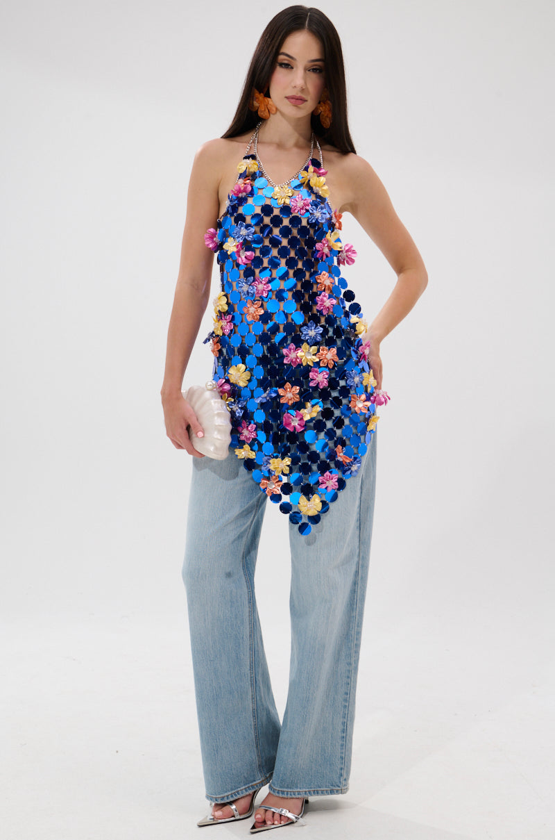 A woman stands against a white background wearing wide-leg jeans, silver heels, the SUMMER DAYS CHAIN TOP—a cobalt blue sequined halter with colorful flowers—large orange earrings, and holding a white textured clutch.
