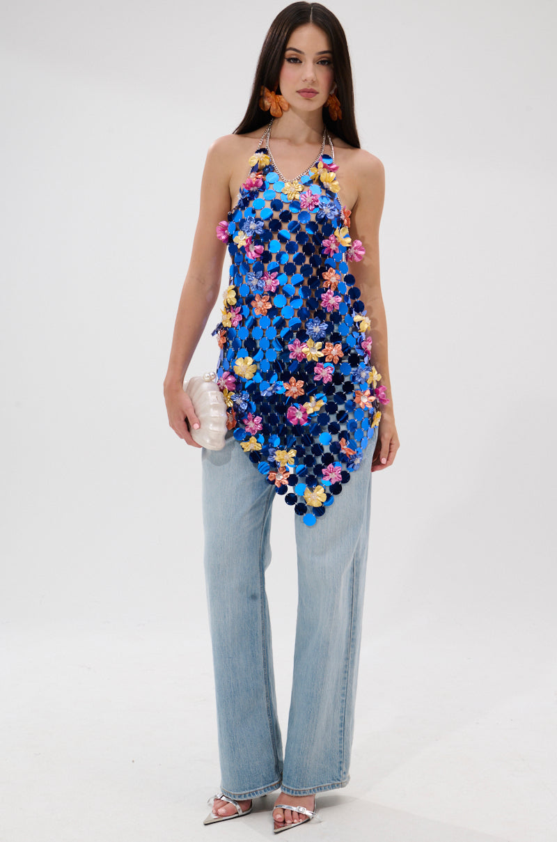 A woman wears the SUMMER DAYS CHAIN TOP, a cobalt blue top with colorful floral details, paired with light-wash wide-leg jeans and a white clutch, posing against a plain white background.