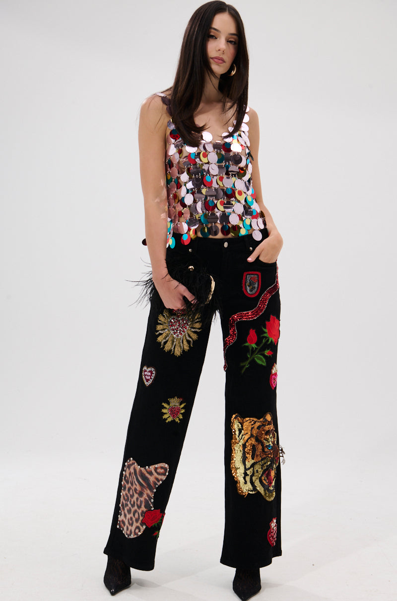 Against a plain backdrop, a woman with long straight hair models the DANCING QUEEN EMBELLISHED TOP—sleeveless and covered in large, colorful sequins—paired with black embroidered pants. She holds a black clutch.