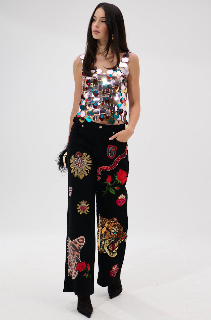 A woman stands against a white background wearing the DANCING QUEEN EMBELLISHED TOP with large metallic sequins, paired with black wide-leg pants featuring tiger and floral patches along with other bold designs.