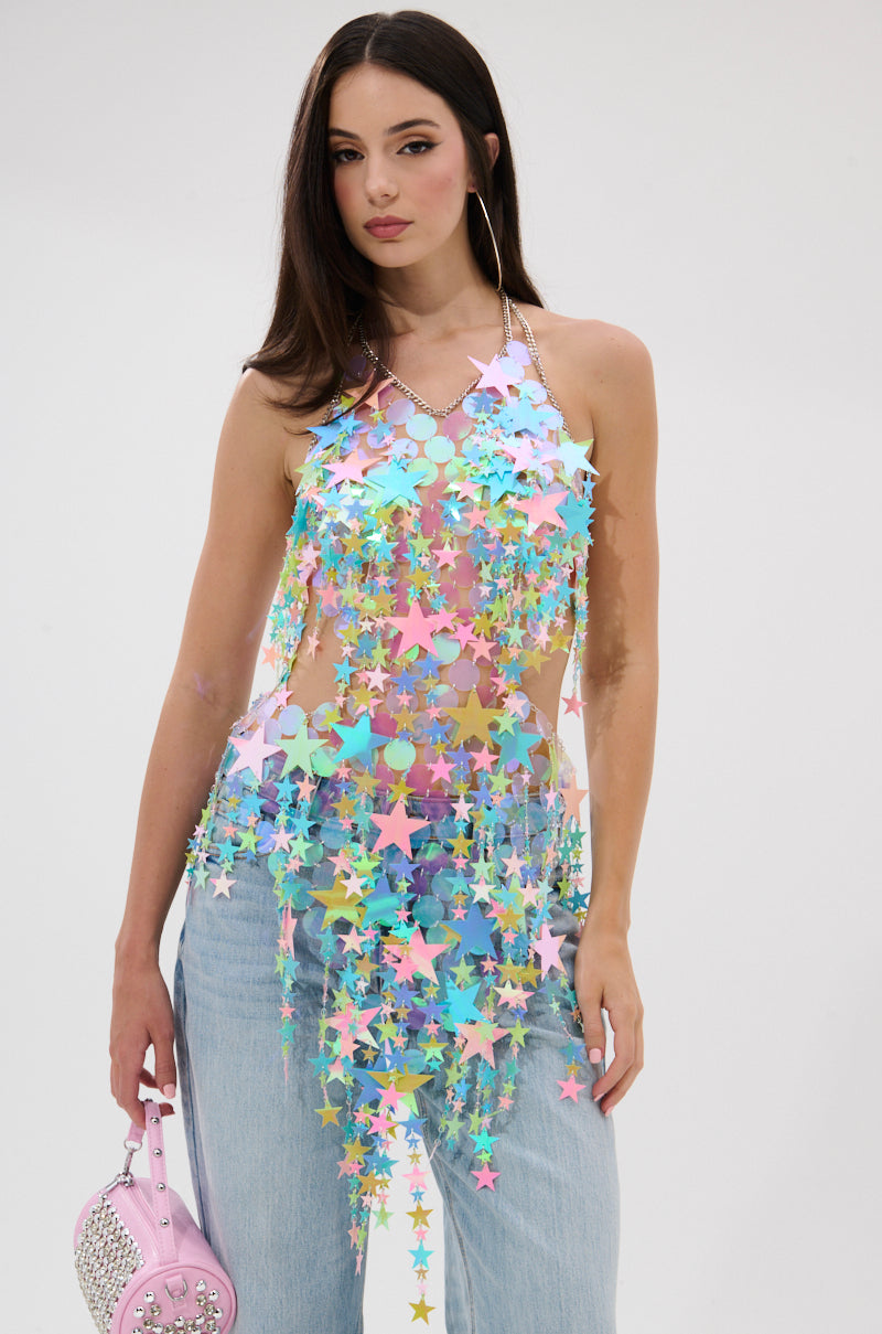 A woman with long dark hair wears the STAR QUEEN BLOUSE, a sheer top with colorful iridescent stars, over light blue jeans. She holds a light pink studded handbag against a plain white background.