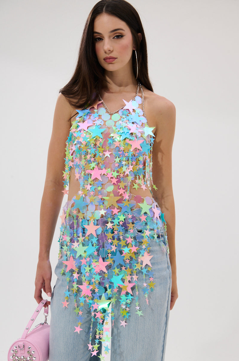 A woman with long dark hair models the STAR QUEEN BLOUSE, a sheer top adorned with colorful iridescent star sequins and pastel charms. She pairs it with light blue jeans, holding a pink purse against a plain white background.