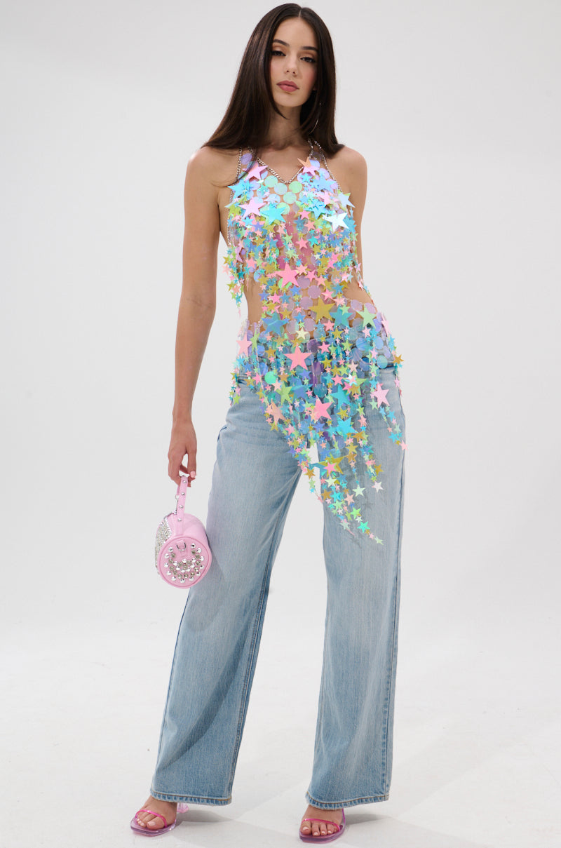 A woman stands against a white background in light blue wide-leg jeans and the STAR QUEEN BLOUSE—a sheer, asymmetrical top with colorful star-shaped sequins. She accessorizes with a pink handbag and matching pink heeled sandals.