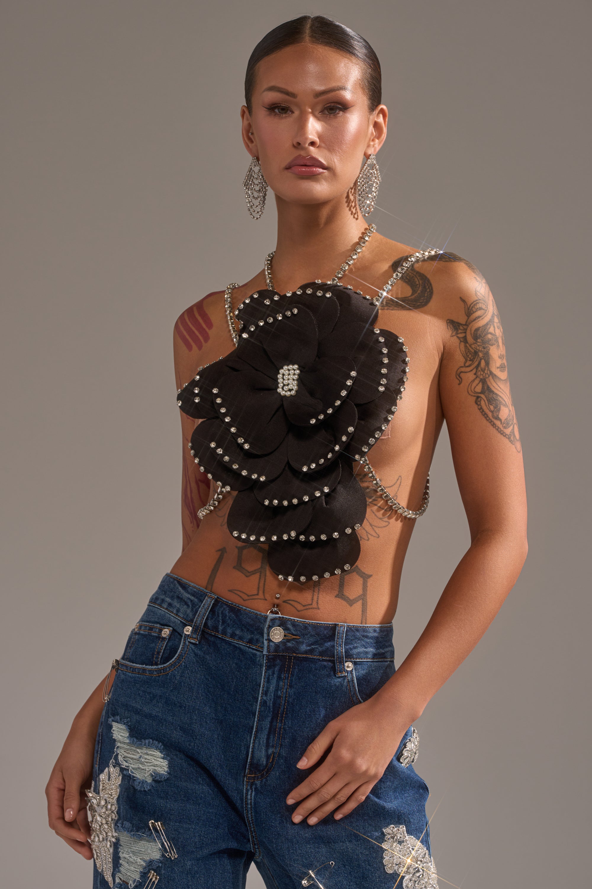 A tattooed woman poses confidently in large hoop earrings, embellished jeans, a striking black floral top with rhinestones, and the LET ME BLOOM BODY CHAIN. The background is simple and neutral. 0