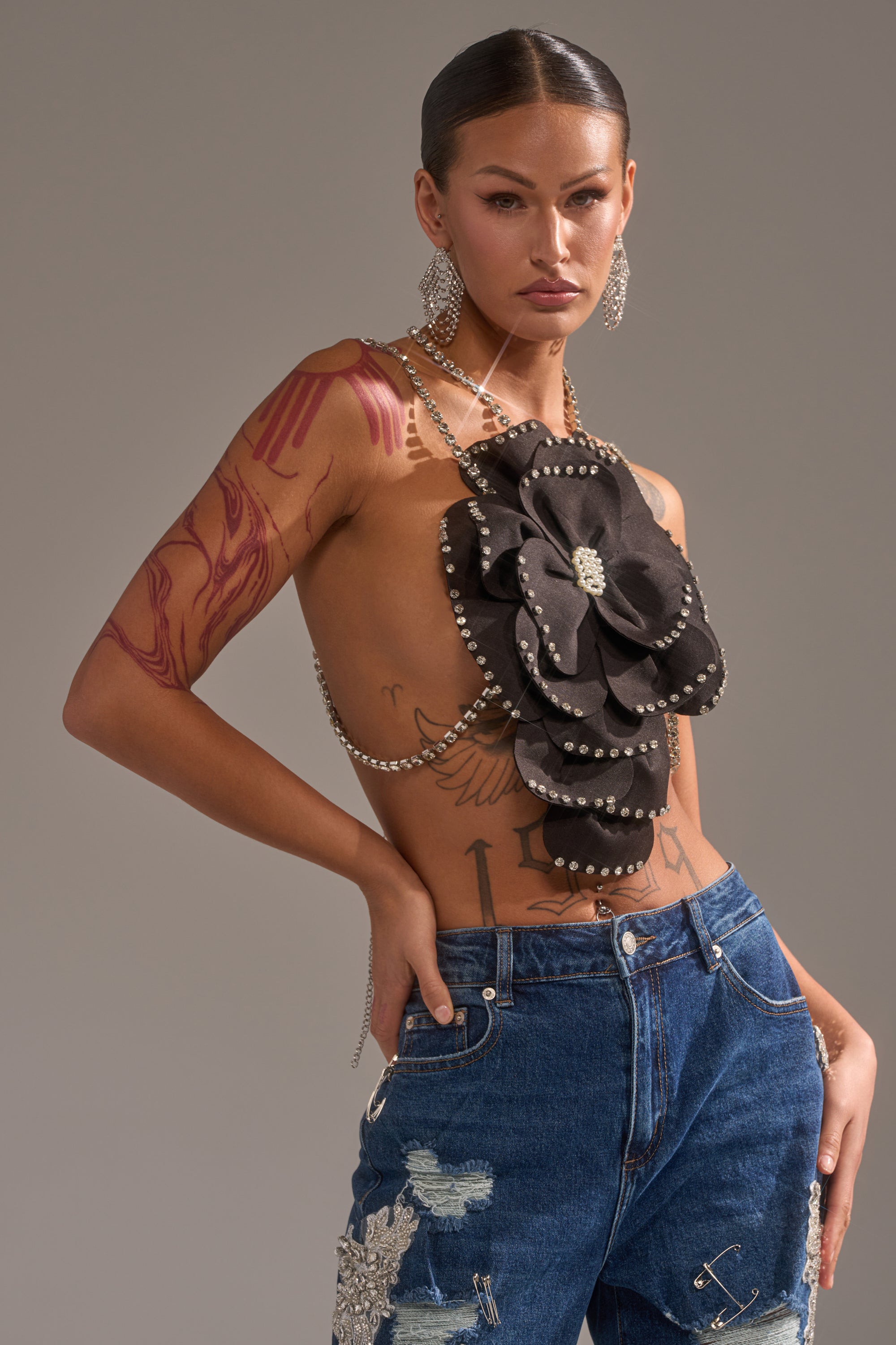 A person poses confidently in blue jeans with patches and a black flower top adorned with pearls, chains, and the LET ME BLOOM BODY CHAIN. Statement earrings complete the look, with visible tattoos on their arm and torso. 1