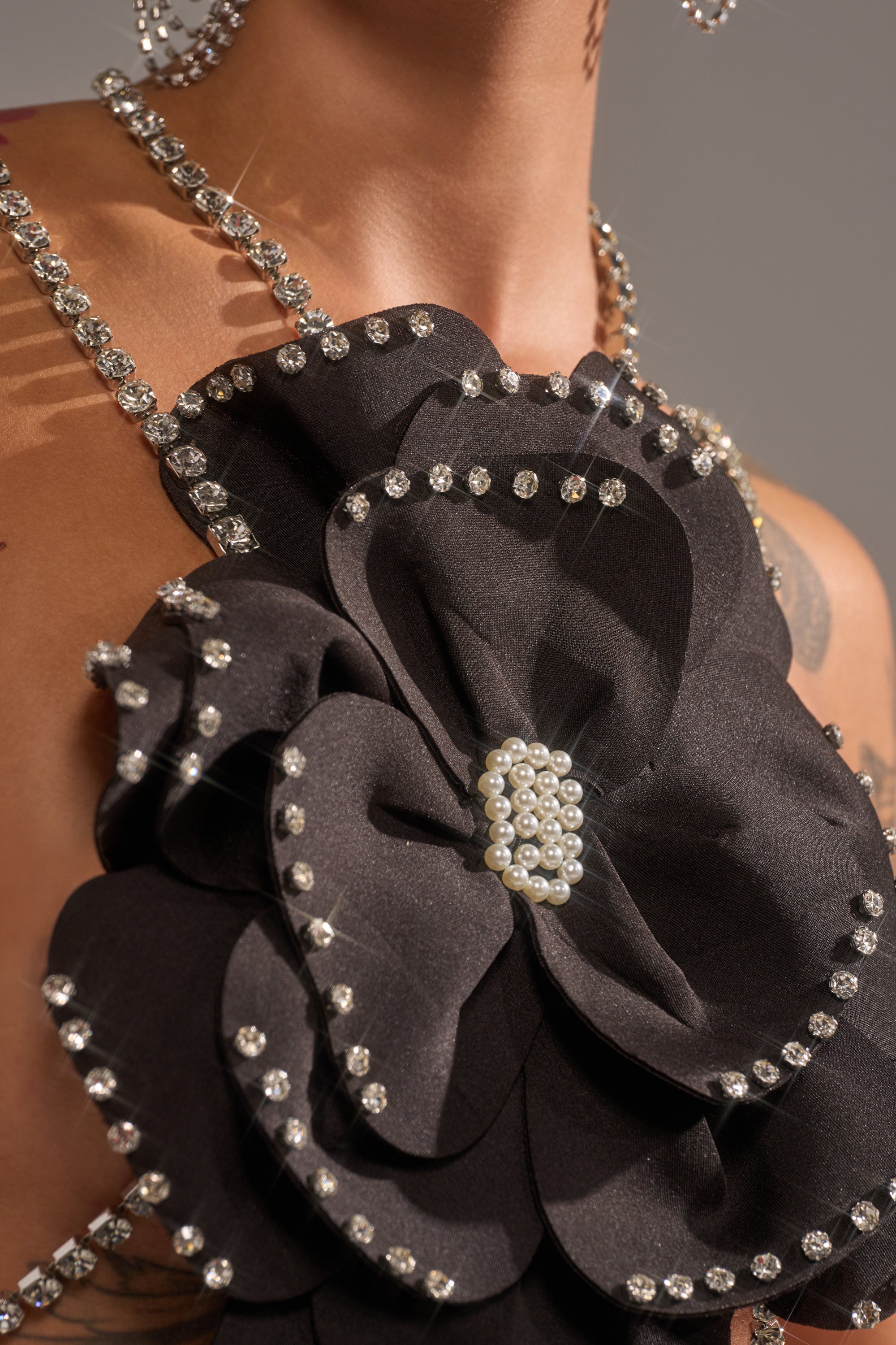 A close-up of a person wearing the LET ME BLOOM BODY CHAIN, featuring a black fabric flower with rhinestones and a pearl center, draped elegantly around their shoulders. 5