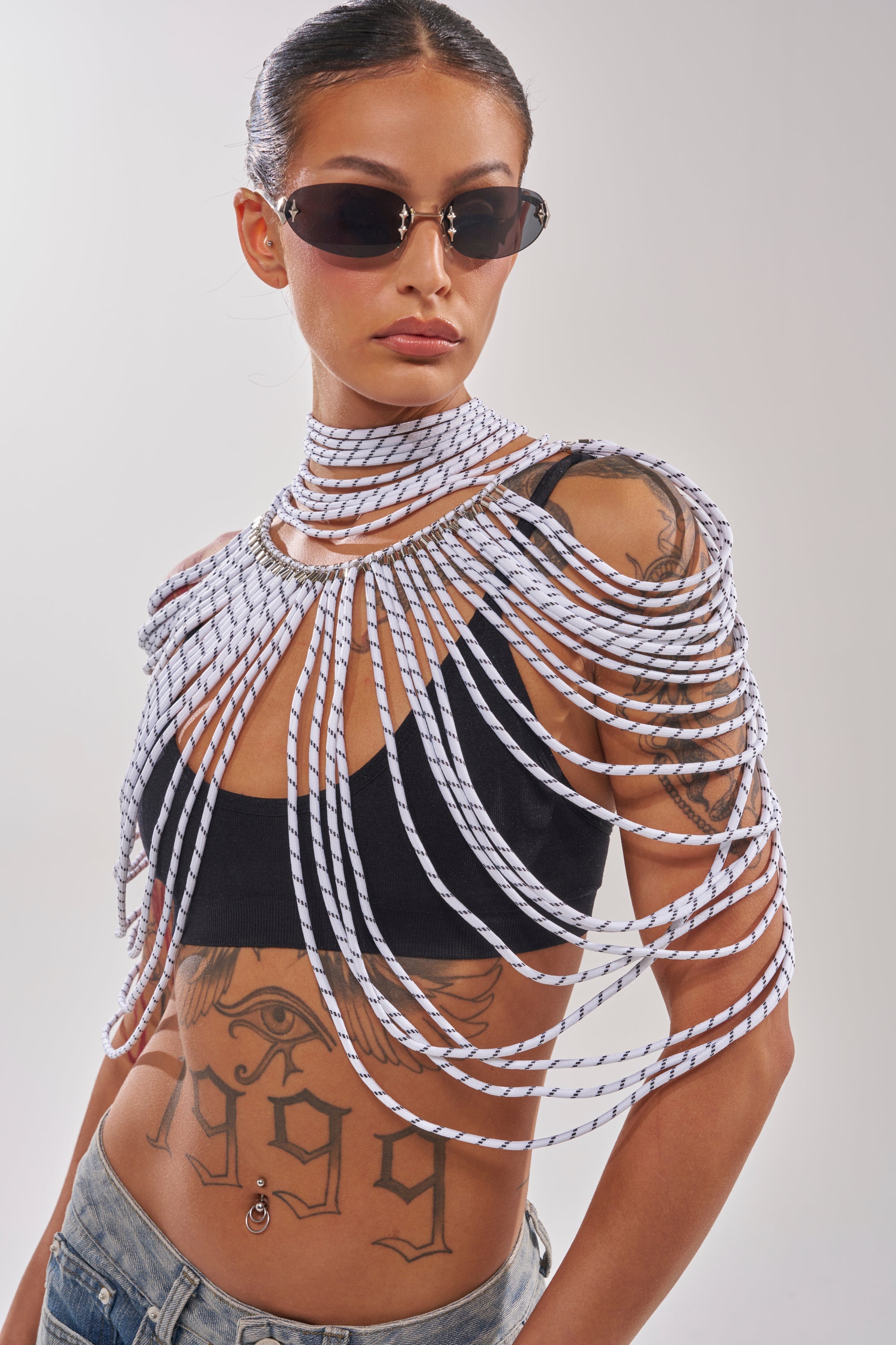 A person wears sunglasses, a black crop top, light-wash jeans, and the COURTSIDE BODY NECKLACE draped over their shoulders and chest. Prominent tattoos are visible on their torso. 1