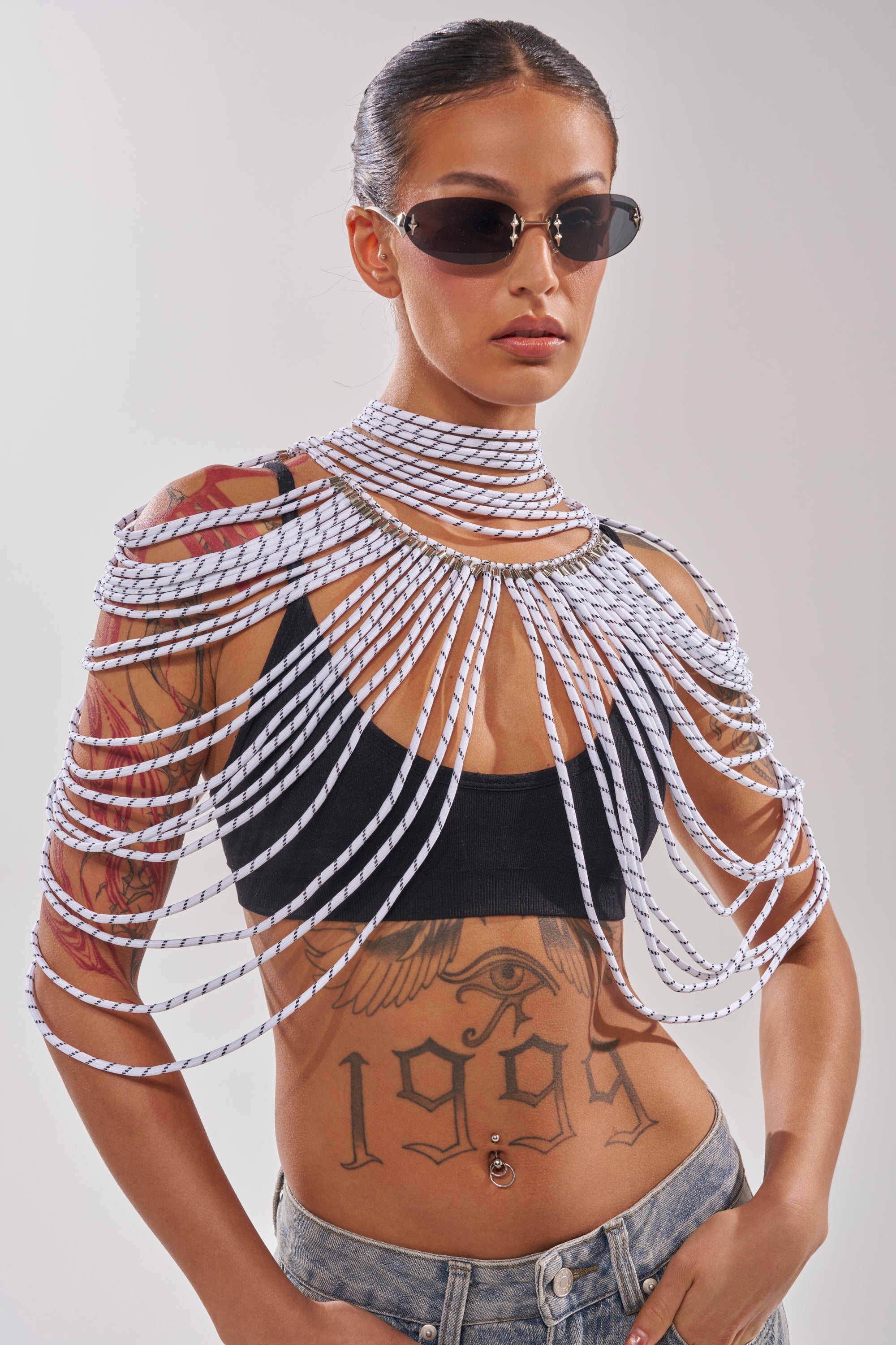 A woman wears sunglasses, a black crop top, and the COURTSIDE BODY NECKLACE draped over her shoulders and arms. She has a torso tattoo and stands against a plain background. 2