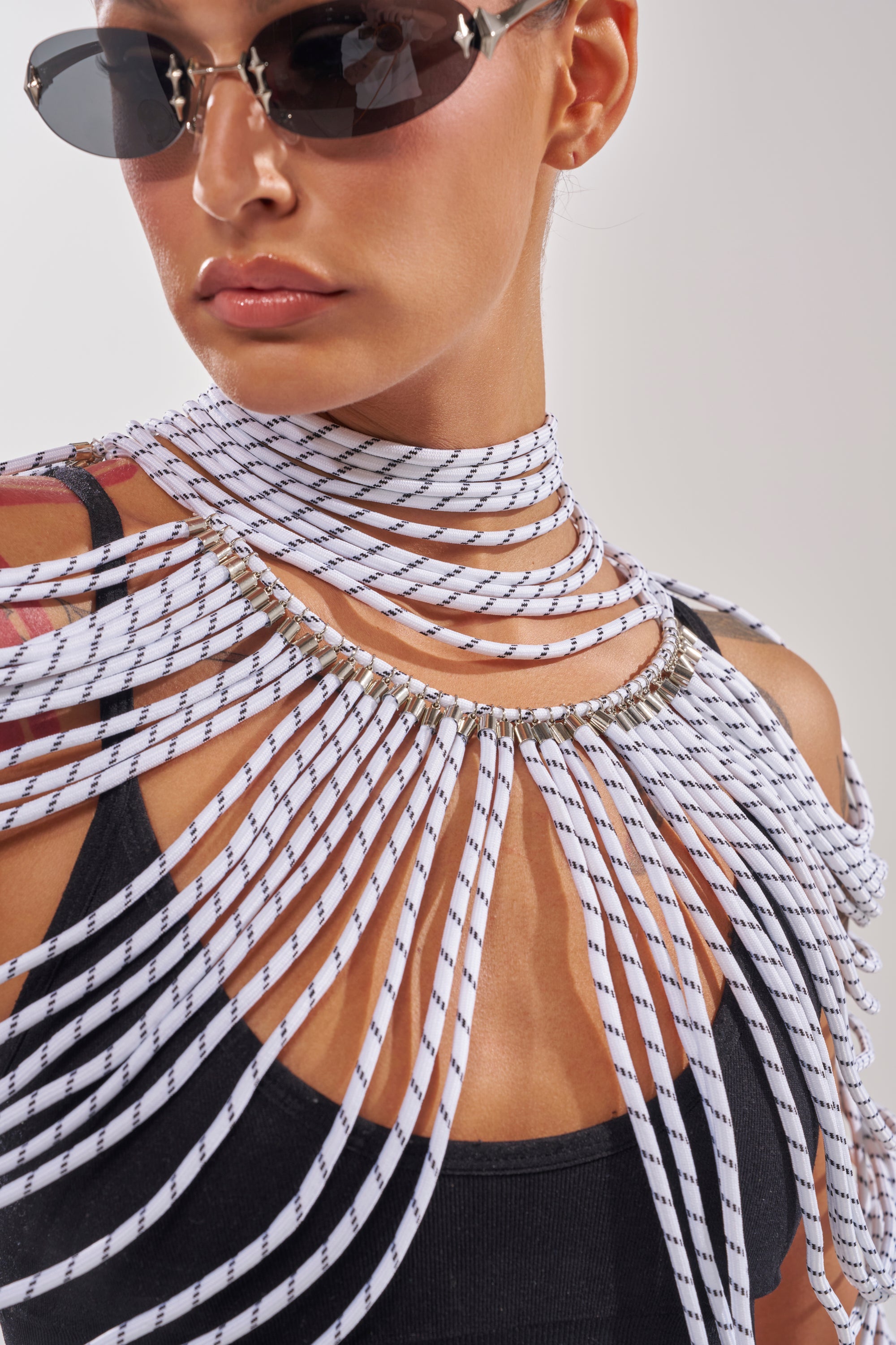 Modeling the COURTSIDE BODY NECKLACE, a woman in sunglasses and a black tank top displays its dramatic, avant-garde design—multiple sculptural white cords with black stripes fanned around her neck and shoulders. 3