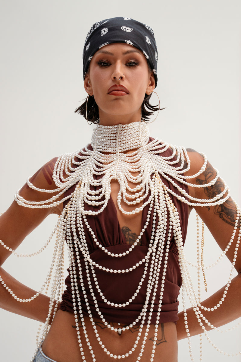 A plain background highlights a person in a black bandana wearing the FEELIN DEMURE BODY CHAIN, an intricate faux pearl necklace covering the shoulders and chest, draped over a sleeveless maroon top. 2