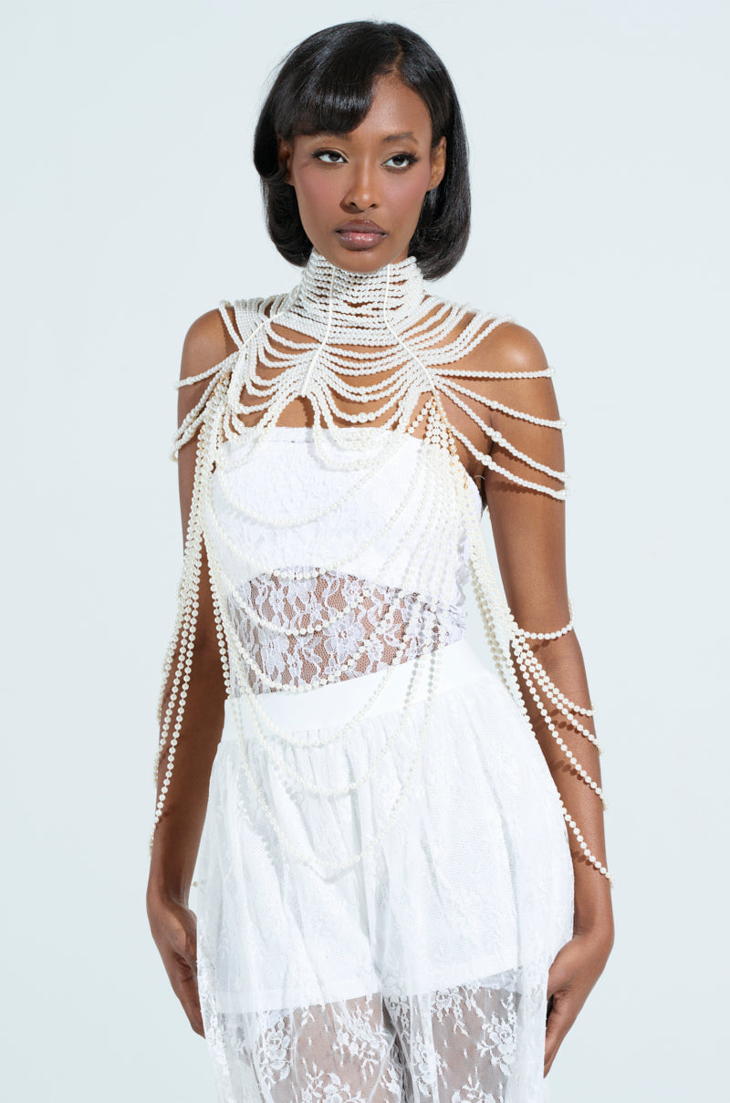 A person in a white lace dress wears the FEELIN DEMURE BODY CHAIN, embellished with faux pearls and an adjustable clasp. They have a bob hairstyle, standing against a light neutral background. 8