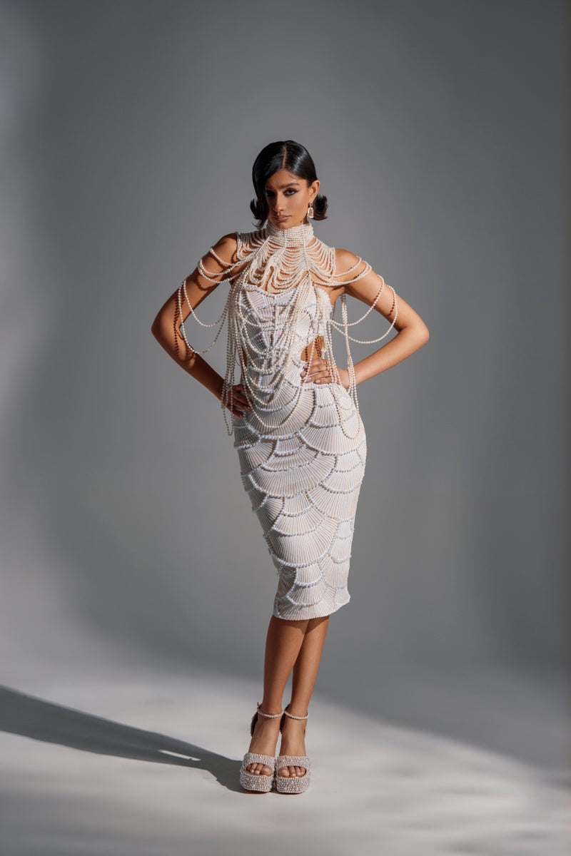 A woman stands confidently in a studio, wearing a fitted white skirt and crop top with scalloped details, accented by the FEELIN DEMURE BODY CHAIN draped over her outfit. Her sleek, side-parted hair and matching high-heeled sandals complete the look. 6