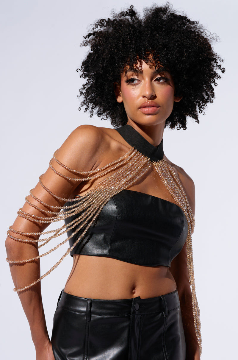 A woman with curly hair wears a black leather tube top paired with high-waisted leather pants. Over her outfit, she elegantly drapes the BELLE OF THE BALL CRYSTAL BODY CHAIN across one shoulder and chest. She stands against a white background, looking confidently ahead.