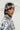 A woman in a shiny metallic outfit adorned with large sequins wears a CLEAR TO ME DRIPPY BUCKET HAT and silver earrings, her hair styled elegantly. Adding a touch of glamour, translucent bead accents complete the look against a plain, light background.