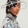 A woman in a shiny metallic outfit adorned with large sequins wears a CLEAR TO ME DRIPPY BUCKET HAT and silver earrings, her hair styled elegantly. Adding a touch of glamour, translucent bead accents complete the look against a plain, light background.