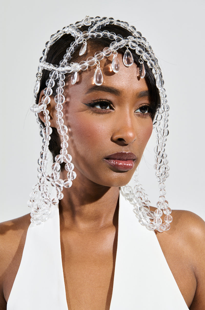 A person wearing a white outfit showcases the CLEAR TO YOU DRIPPY HEAD PIECE, a striking headdress adorned with translucent bead embellishments that cascade elegantly over their forehead and shoulders. The plain background accentuates the intricate design of this headpiece. 0