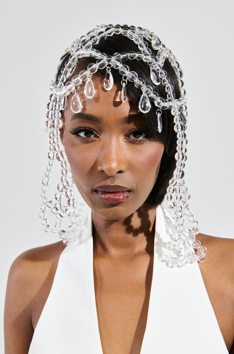 A person wearing a white outfit is adorned with the CLEAR TO YOU DRIPPY HEAD PIECE, which features translucent bead embellishments, including teardrop shapes. The beads elegantly drape over their forehead and shoulders against a neutral background. 2