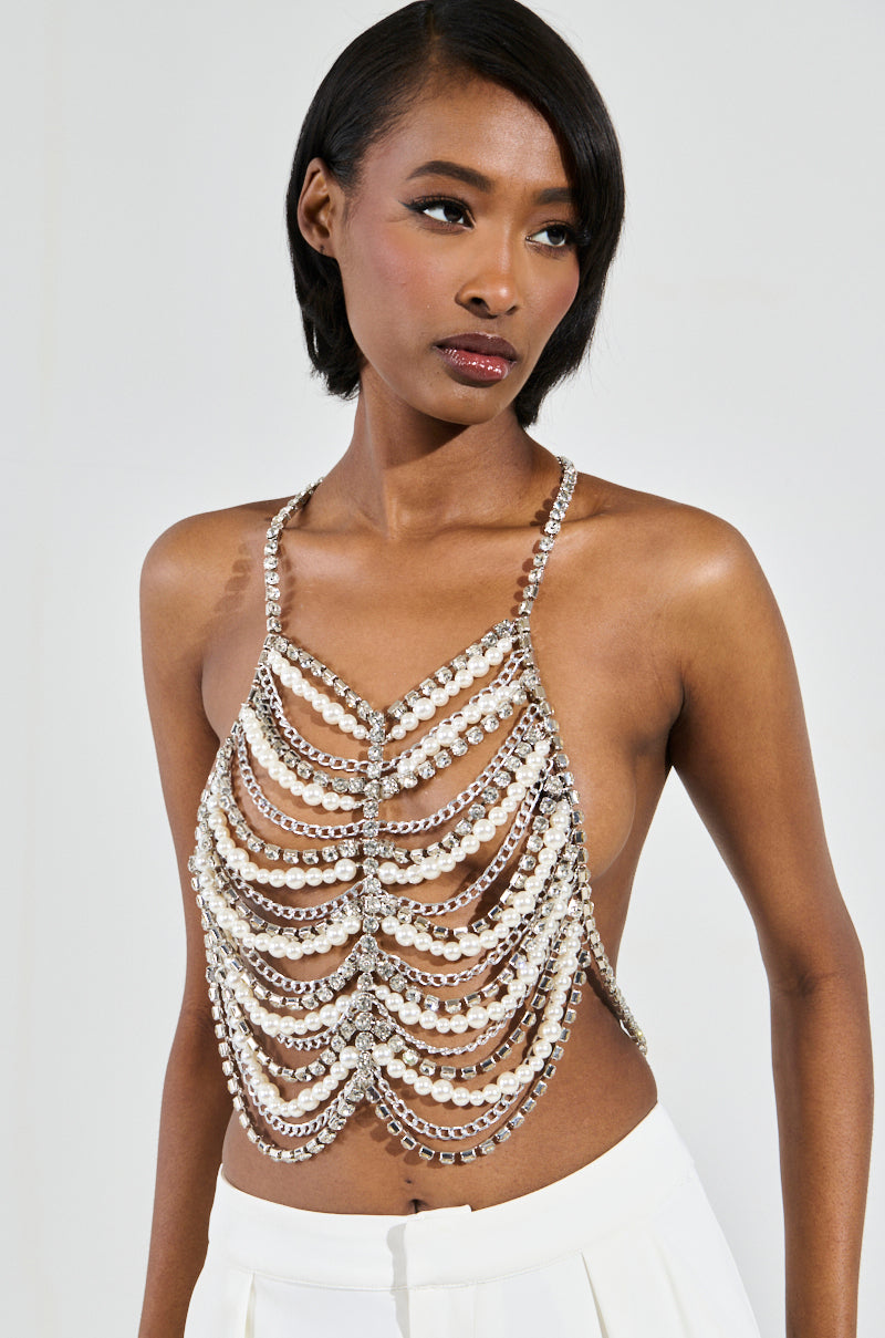 A person poses elegantly in a halter neck outfit featuring the EVA Pearl and Rhinestone Body Chain, paired with white pants, against a plain background.