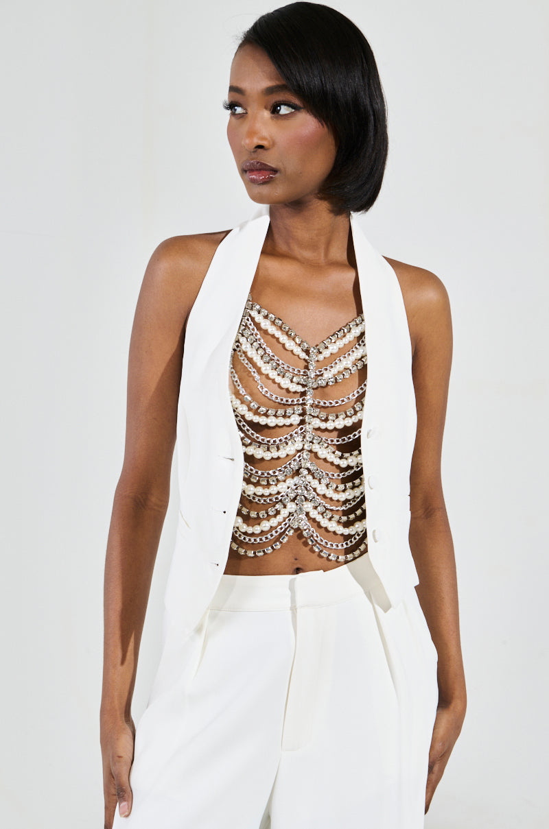 A woman in a chic white ensemble sports a sleeveless tailored jacket and wide-legged pants, revealing the EVA PEARL AND RHINESTONE BODY CHAIN beneath. Her confident pose is complemented by sleek, side-parted hair.