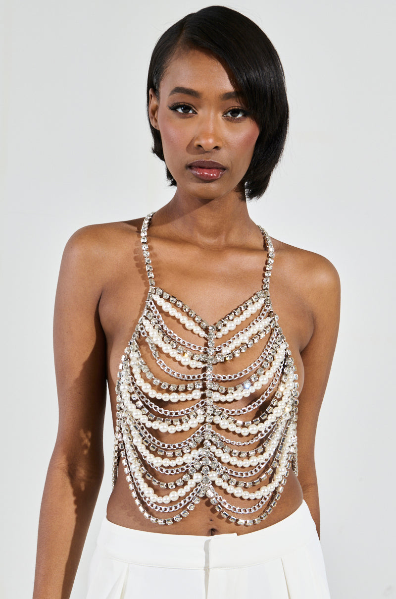 A person with a short hairstyle is wearing the EVA Pearl and Rhinestone Body Chain styled as a halter neck top, paired with white pants. They stand against a plain backdrop, looking directly at the camera with a neutral expression.
