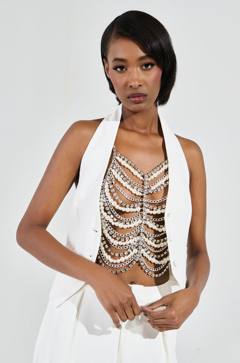A person poses confidently in a stylish white outfit, showcasing a sleeveless jacket and the EVA Pearl and Rhinestone Body Chain. The plain white background highlights their fashion-forward attire and poised demeanor.