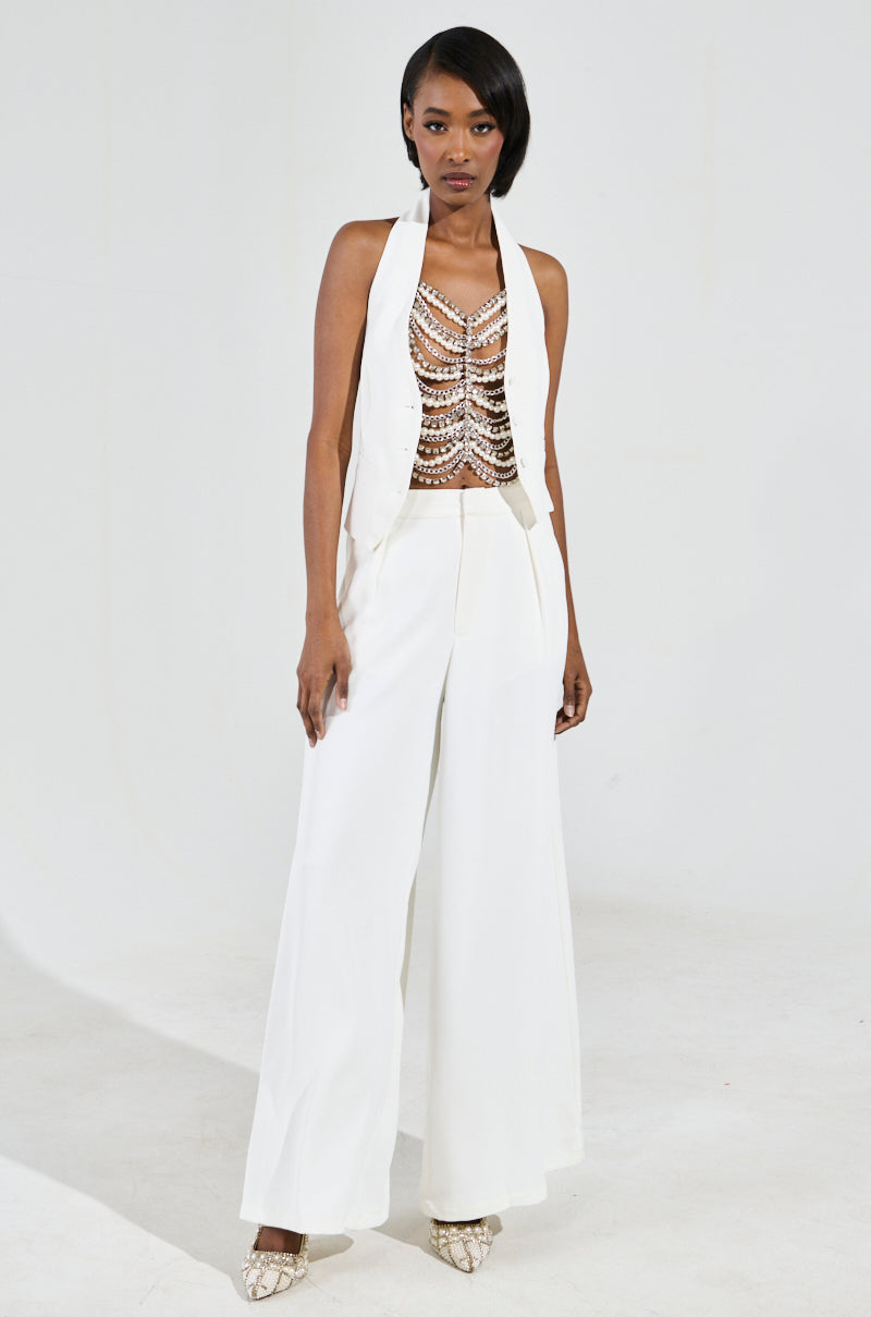 A woman poses confidently against a plain backdrop in a chic white pantsuit featuring a sleeveless jacket and a beaded, patterned halter top. She accessorizes with the EVA Pearl and Rhinestone Body Chain, which complements her short, dark hair beautifully.