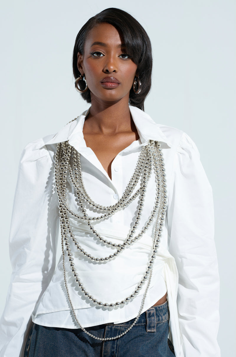 A person in a stylish white blouse wears the ADORN ME LAYERED NECKLACE with metallic silver beads draped across the chest, accessorized with hoop earrings. They pair it with blue jeans and stand against a plain light background. 1