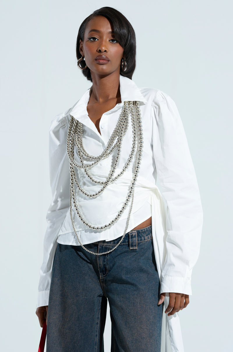 A person in a white button-up shirt accessorized with the ADORN ME LAYERED NECKLACE and statement jewelry stands against a plain background. They also wear blue jeans and have short, styled hair. 2