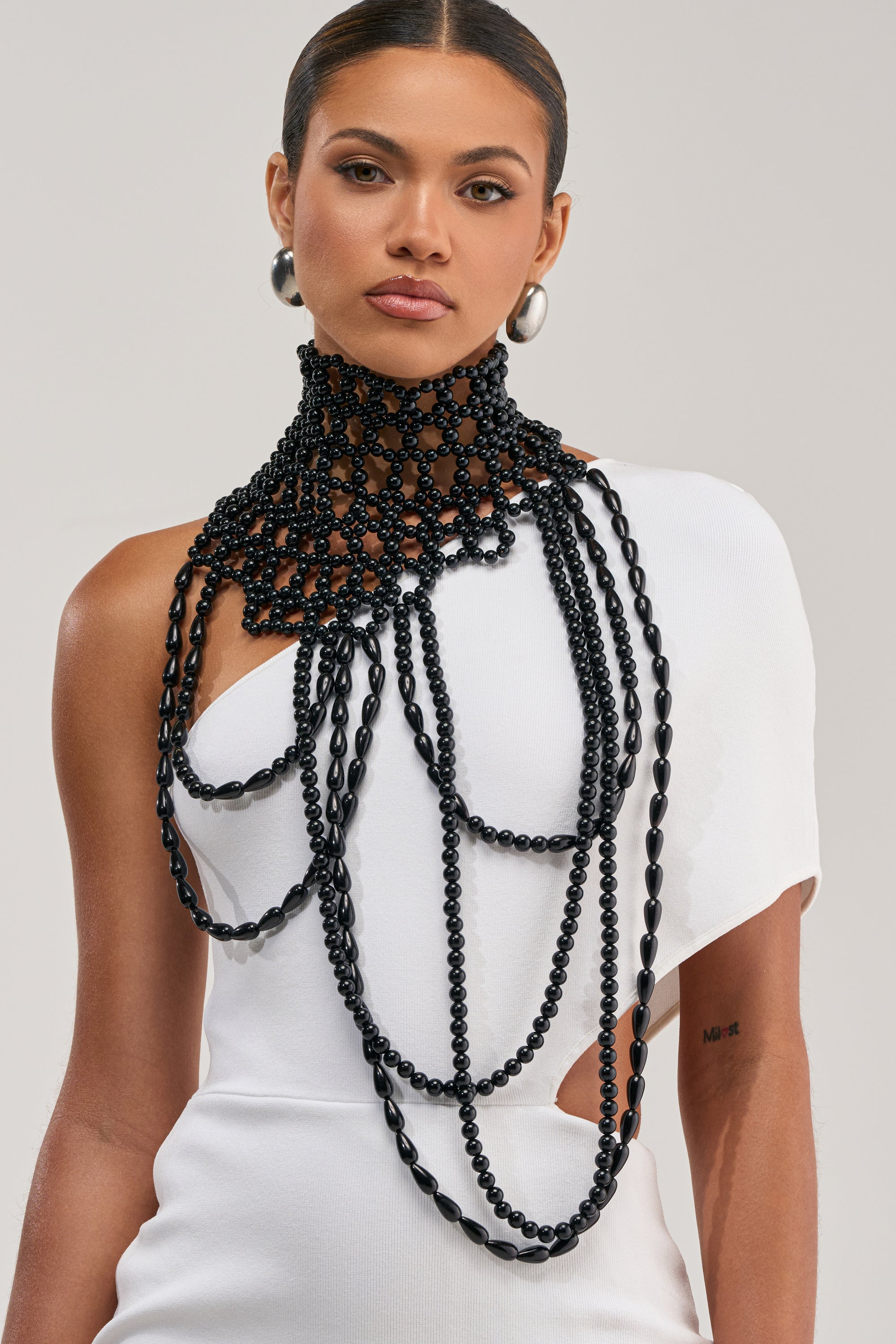A woman in a white one-shoulder dress wears the FASHION WEEK BEADED NECKLACE—a dramatic, layered black beaded piece—paired with large silver hoop earrings. Her hair is neatly styled back as she poses against a plain, light background.