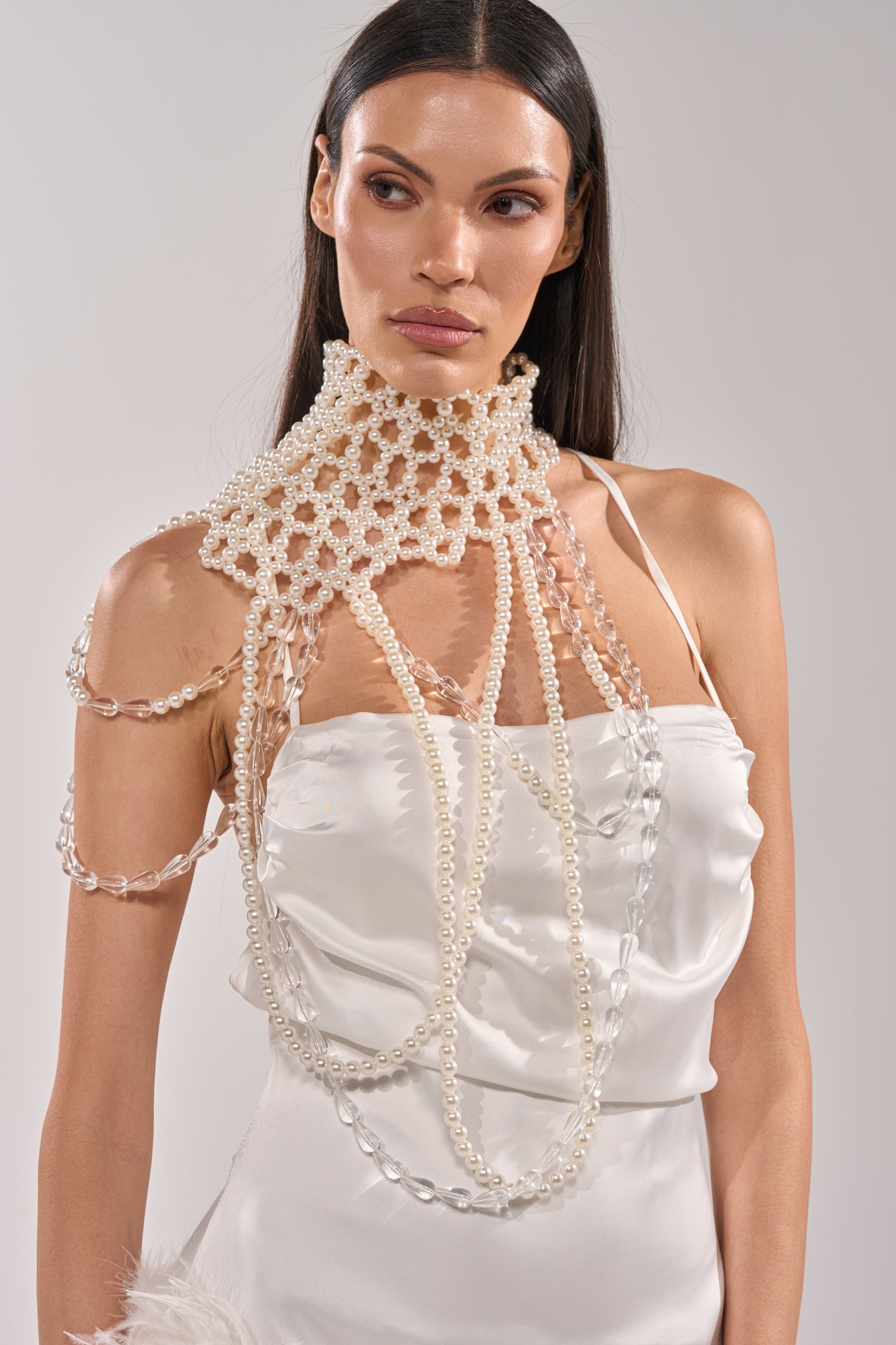 Wearing the ELEGANCE IS ME STATEMENT NECKLACE over a white satin dress, a woman with long dark hair gazes to the side against a light background, highlighting the bold pearl piece draped across her shoulder and chest. 0