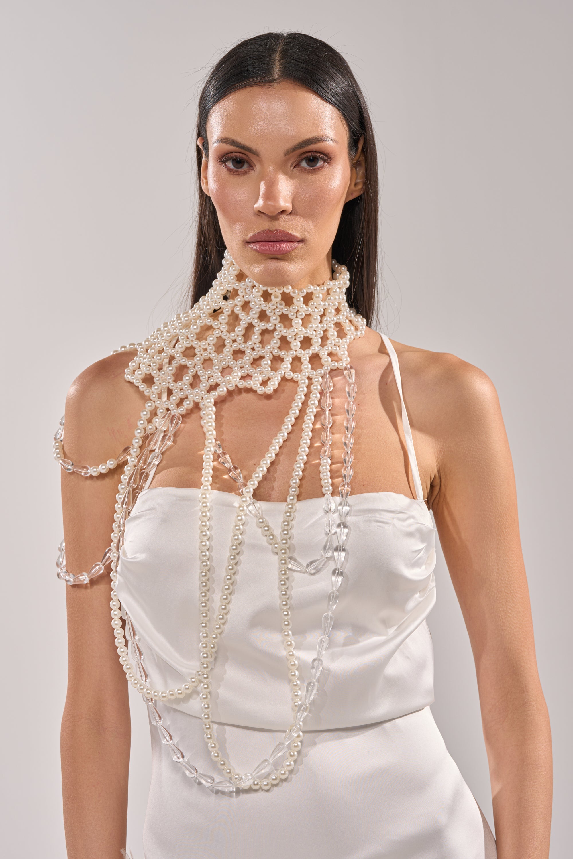 A woman in a white satin dress models the ELEGANCE IS ME STATEMENT NECKLACE, featuring pearls and chains draped around her neck, shoulders, and chest, standing against a plain light background. 1