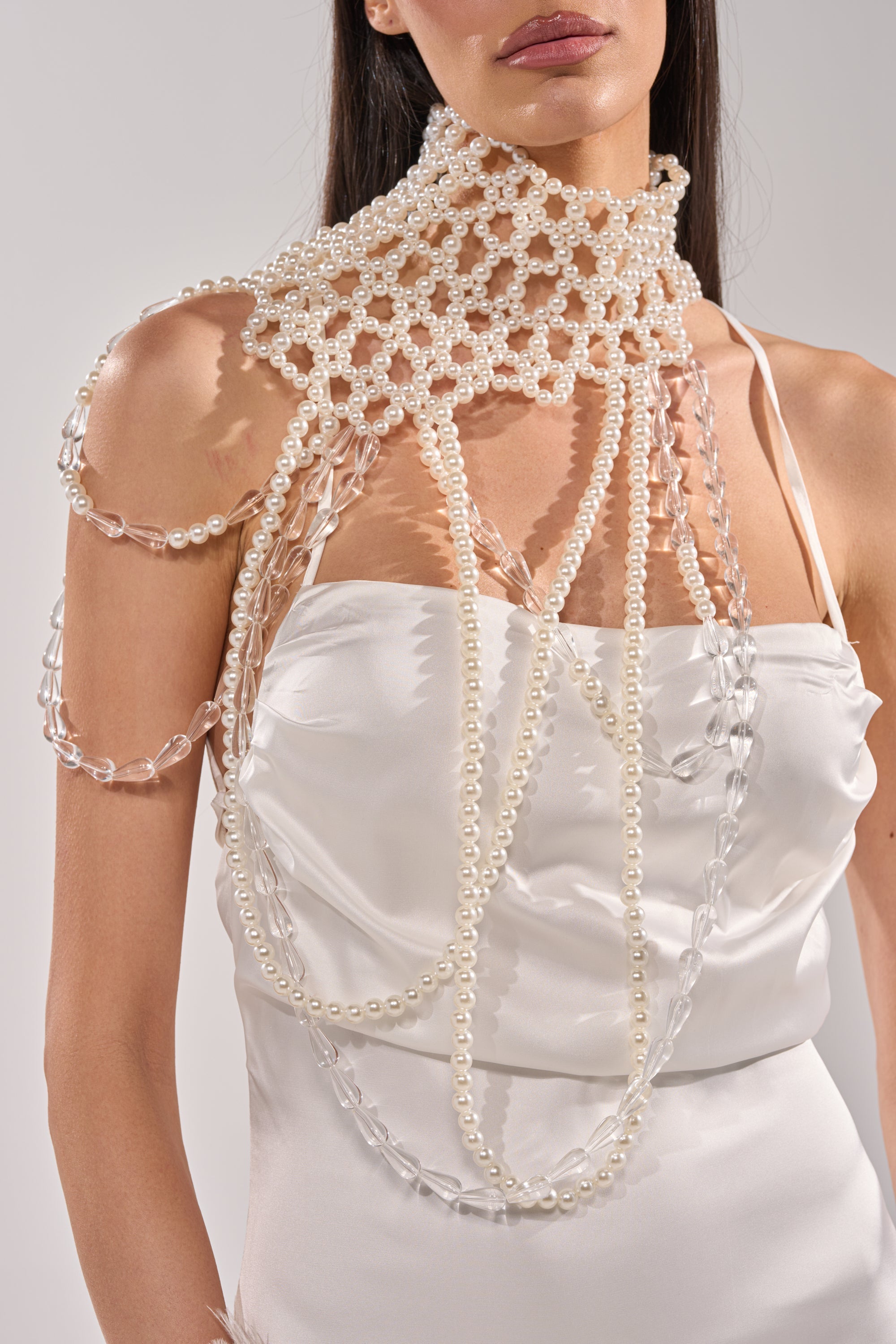 A woman in a white satin dress wears the ELEGANCE IS ME STATEMENT NECKLACE, featuring pearls and clear beads in a net-like design over her neck, shoulders, and upper chest. 2