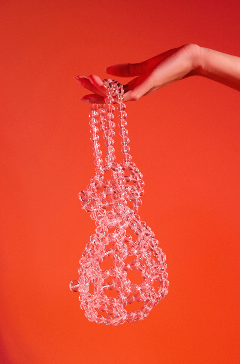 A hand holds the CLEAR VIEW MINI SACK BAG, a transparent handbag crafted from clear, round beads with an intricate net-like design, shimmering against a vivid red-orange background.
