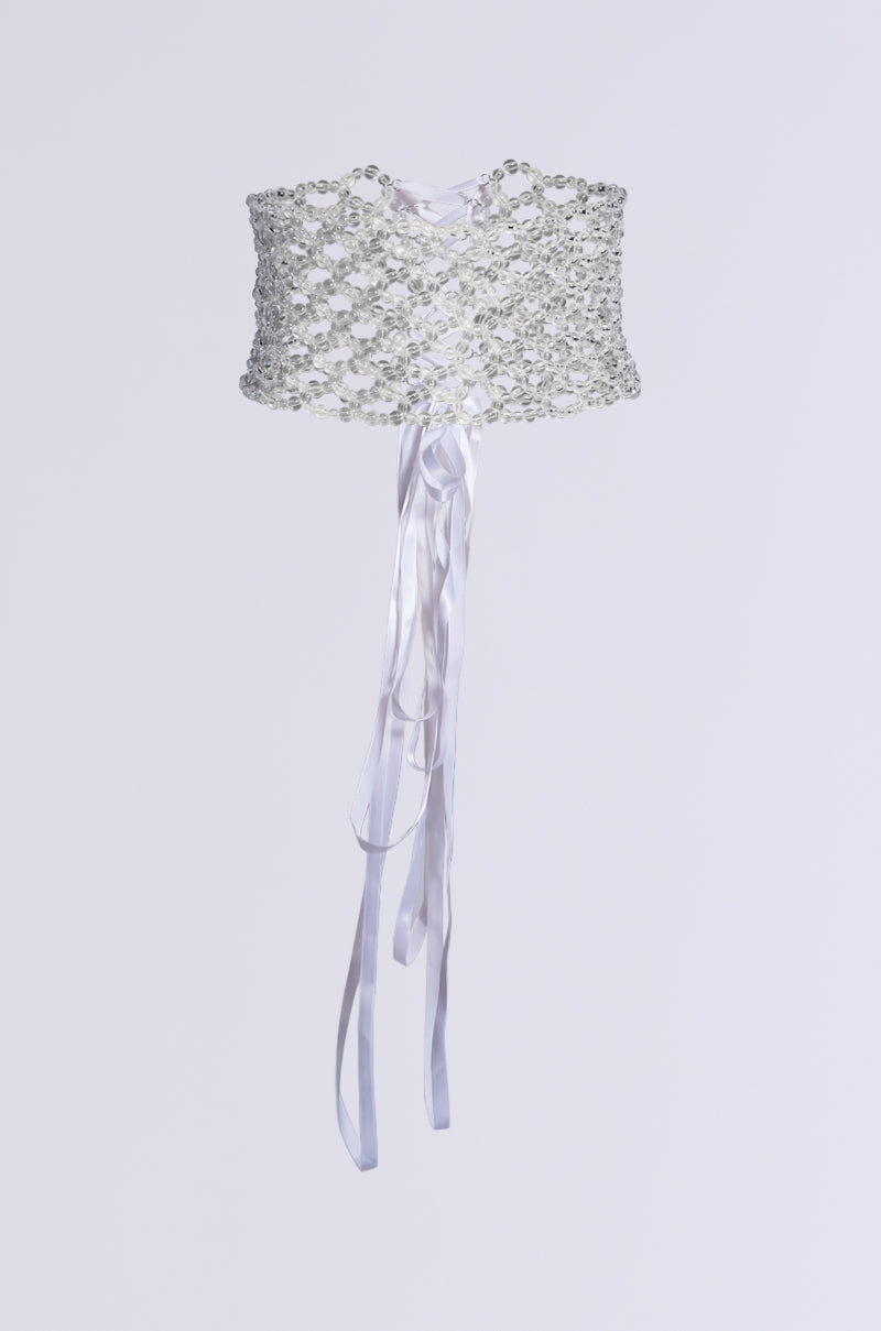 The AKIRA Label presents the CLEAR VIEW BEADED LACEUP BELT, a stunning white corset adorned with sparkling beads and jewels. This elegant accessory, featuring a lace-up design with long satin ribbons, stands out against its plain white backdrop.