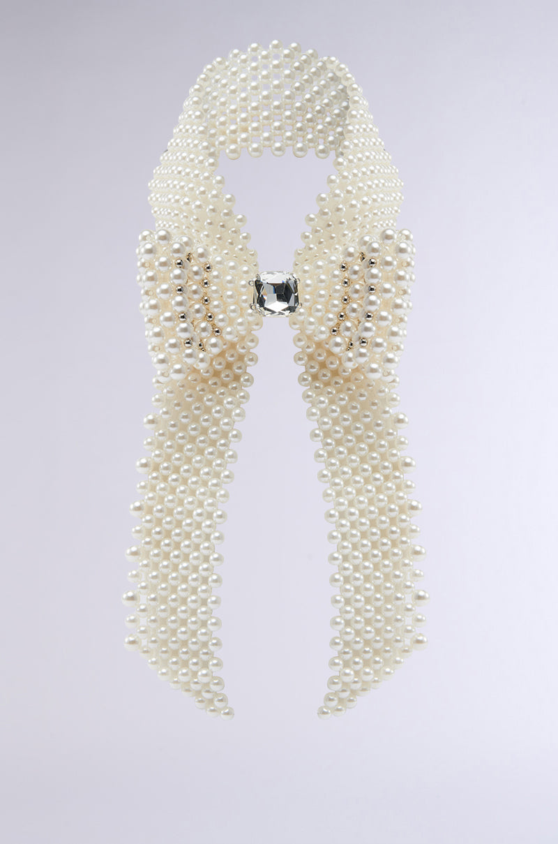 The BUTTONED UP PEARL COLLAR, a bow-shaped necklace with rows of white pearls and a central rectangular crystal, channels vintage charm and makes a standout statement against a light gray background. 6