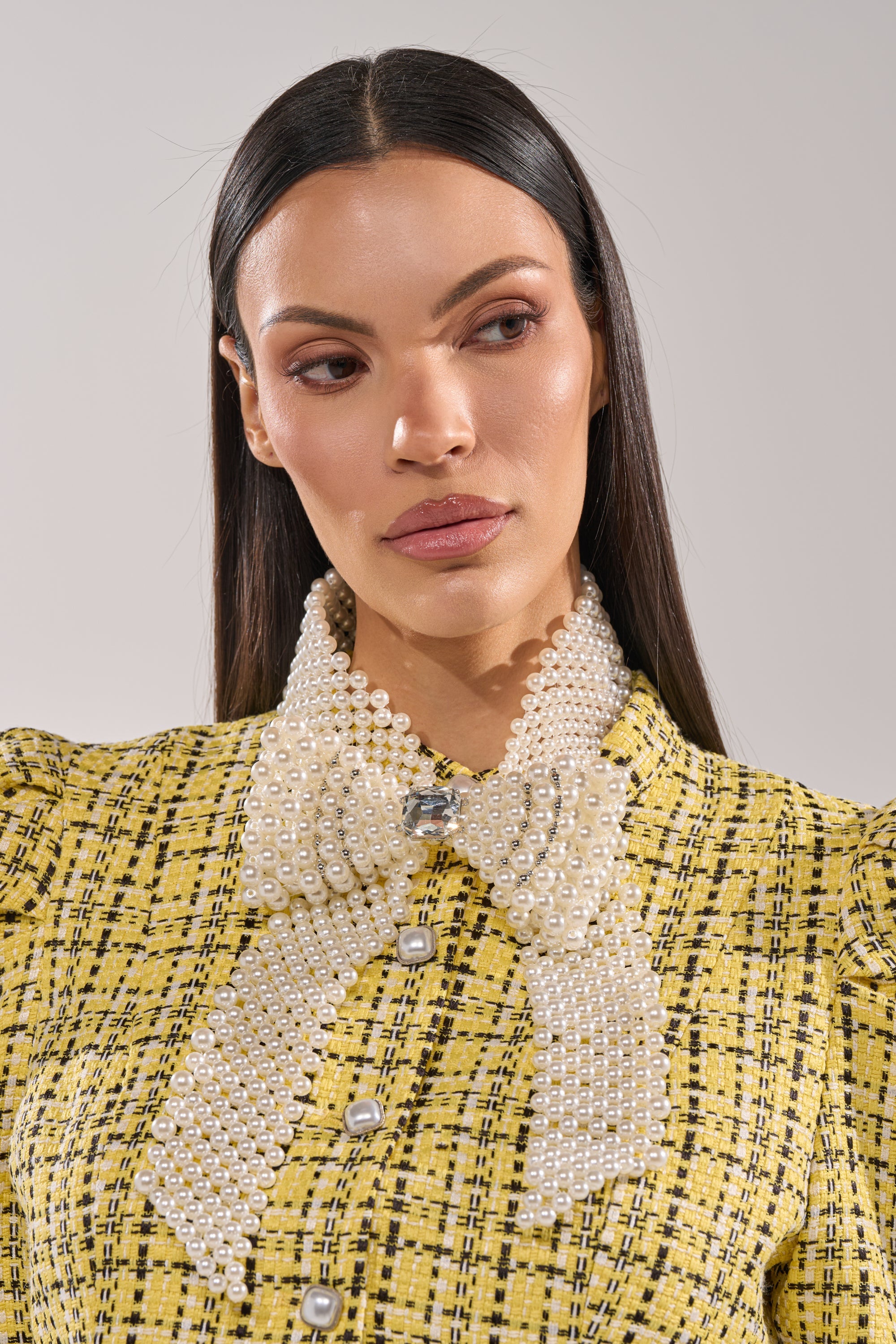 A woman with straight dark hair wears the BUTTONED UP PEARL COLLAR, featuring a yellow textured jacket, pearl buttons, and a vintage-inspired pearl bow collar. She looks slightly to the side against a plain light background. 0