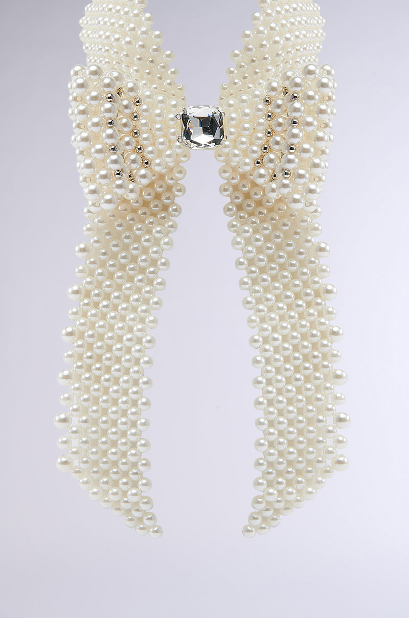 The BUTTONED UP PEARL COLLAR features multiple strands of white pearls forming a decorative bow with a large clear gemstone center, exuding vintage charm and showcased against a light gray backdrop. 7