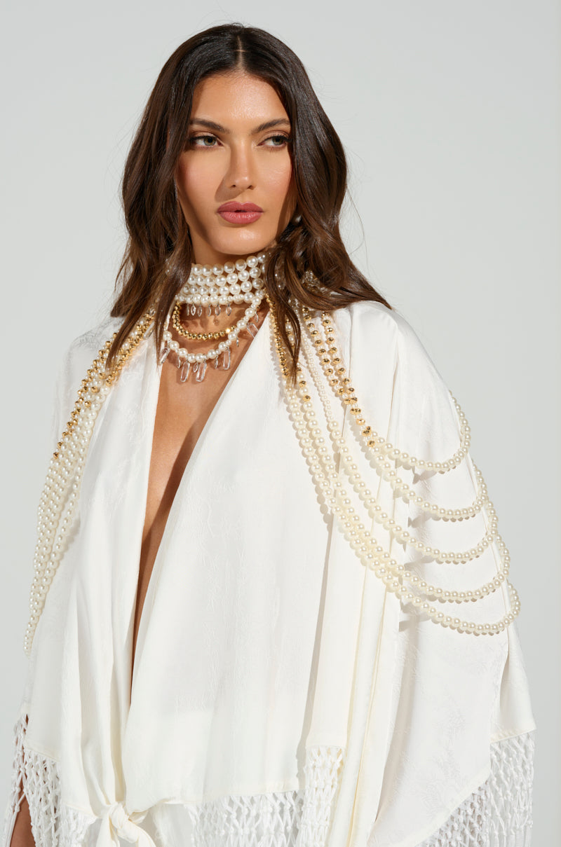A long-haired woman in a textured white outfit with fringe wears the OUT OF YOUR LEAGUE NECKLACE, a layered pearl choker with gold details, against a plain light gray background.