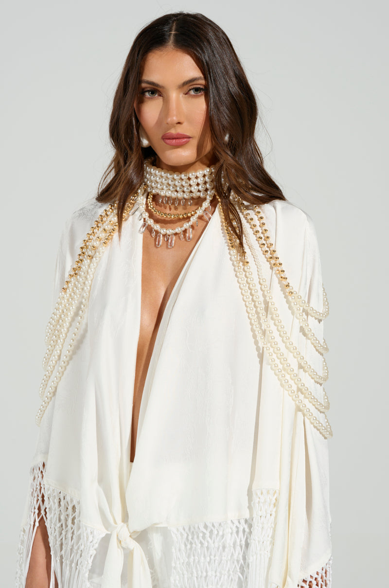 A woman with long brown hair wears a white garment with gold embellishments and a deep neckline. Layered pearl necklaces, including the OUT OF YOUR LEAGUE NECKLACE, adorn her neck, complementing the intricate beading as she stands against a plain background.