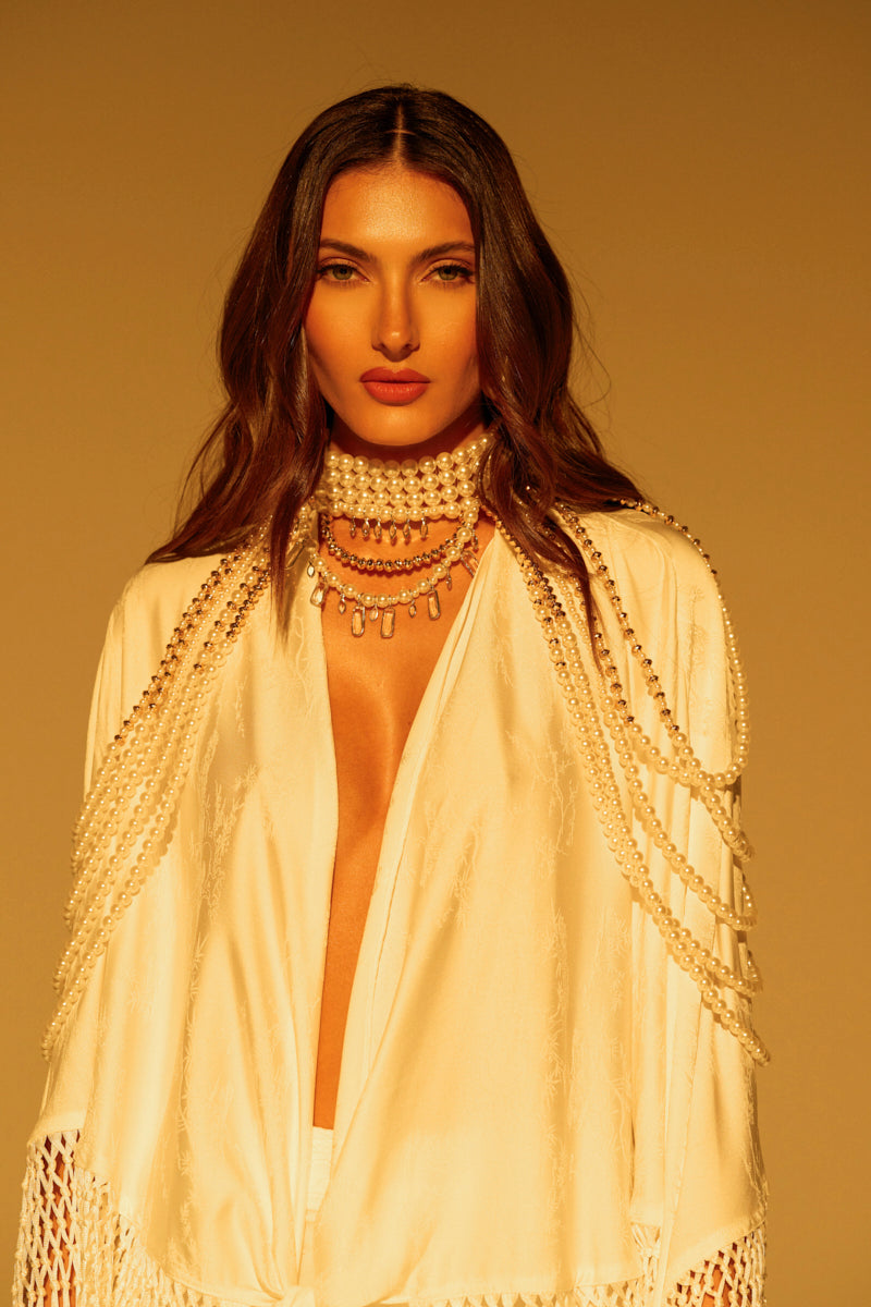 A person with long brown hair wears an elegant, flowing white outfit enhanced by gold embellishments, adorned with the "Out of Your League Necklace" featuring multiple pearl layers. The warm golden hue background amplifies the sophisticated look.