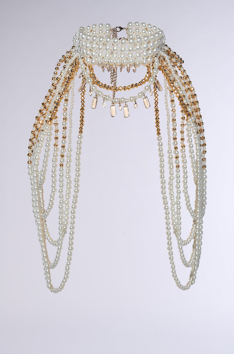 The OUT OF YOUR LEAGUE NECKLACE features an ornate headpiece with a pearl and gold chain circular base, cascading strands, and small gold charms, resembling a pearl choker necklace against a plain backdrop.