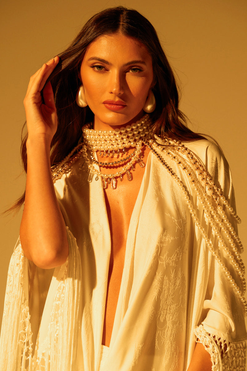 A woman in a white outfit is adorned with the "OUT OF YOUR LEAGUE NECKLACE," featuring a pearl choker and multiple strands of pearls. She has long dark hair, wears large pearl earrings, and poses against a softly lit background that creates a warm ambiance.