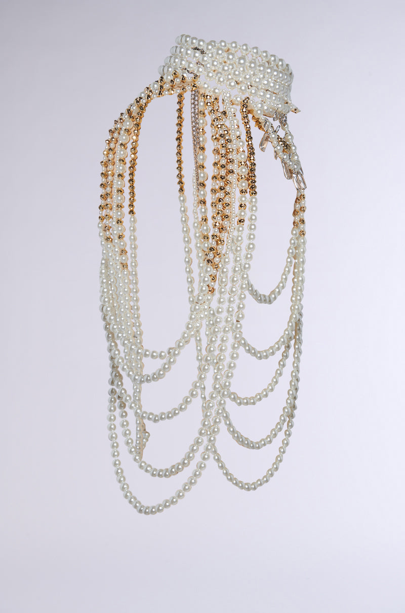 The OUT OF YOUR LEAGUE NECKLACE, a pearl choker adorned with cascading strands of pearls and gold chains, features decorative gemstone pendants. It drapes elegantly against a light background, highlighting its timeless beauty and intricate design.