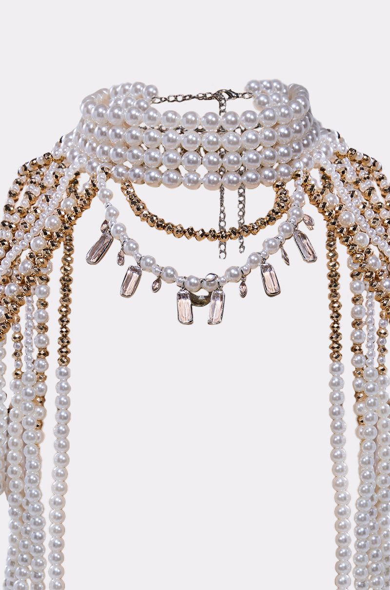 The OUT OF YOUR LEAGUE NECKLACE is a layered choker featuring white pearls, gold beads, and clear crystal pendants in cascading strands, offering an elegant and ornate design.