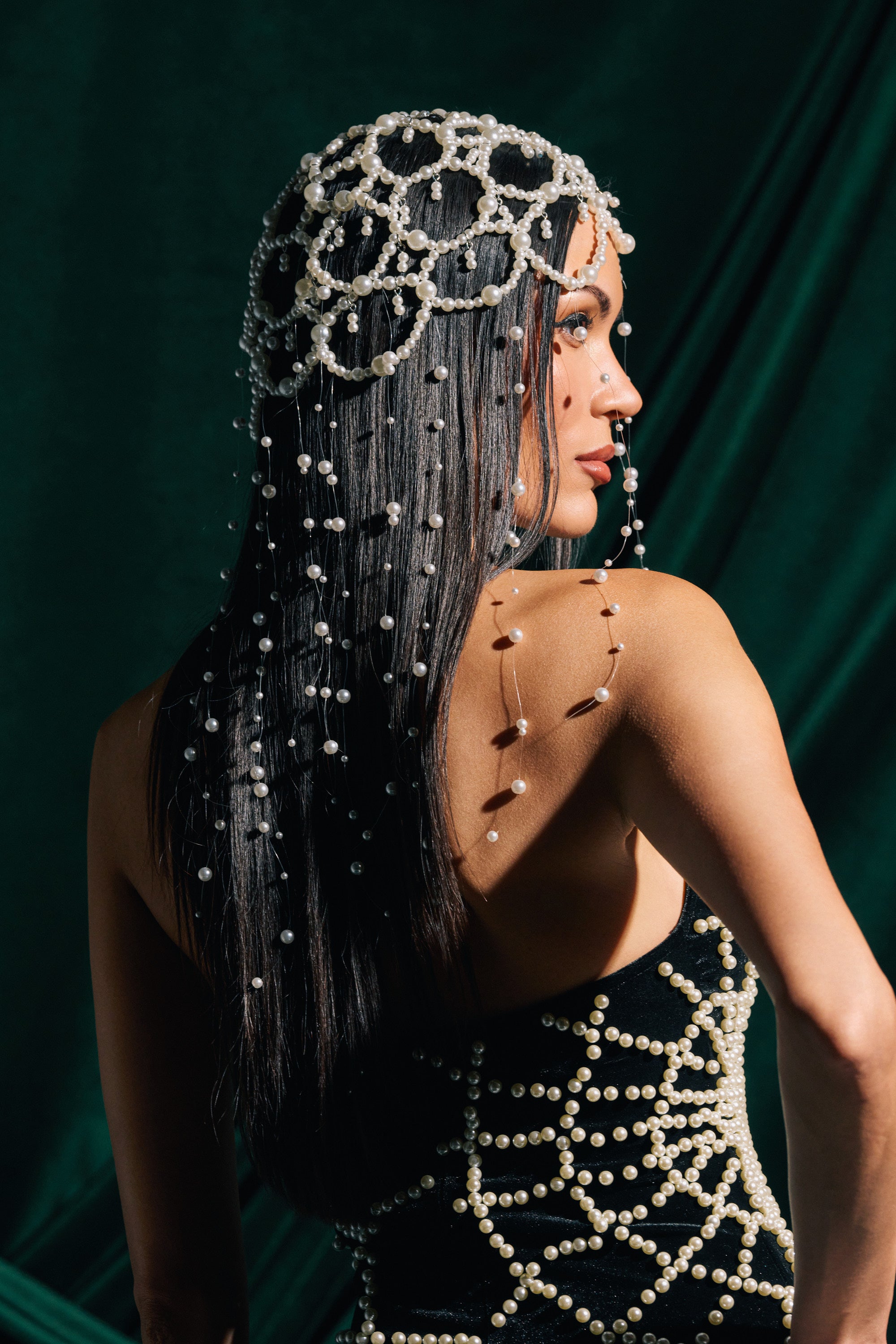 A woman with long dark hair wears the PEARL LOVE HEADPIECE and a black strapless dress with pearl details, standing against a deep green draped backdrop. She faces slightly away, her elegant pearl accessory catching the light as she glances back.
