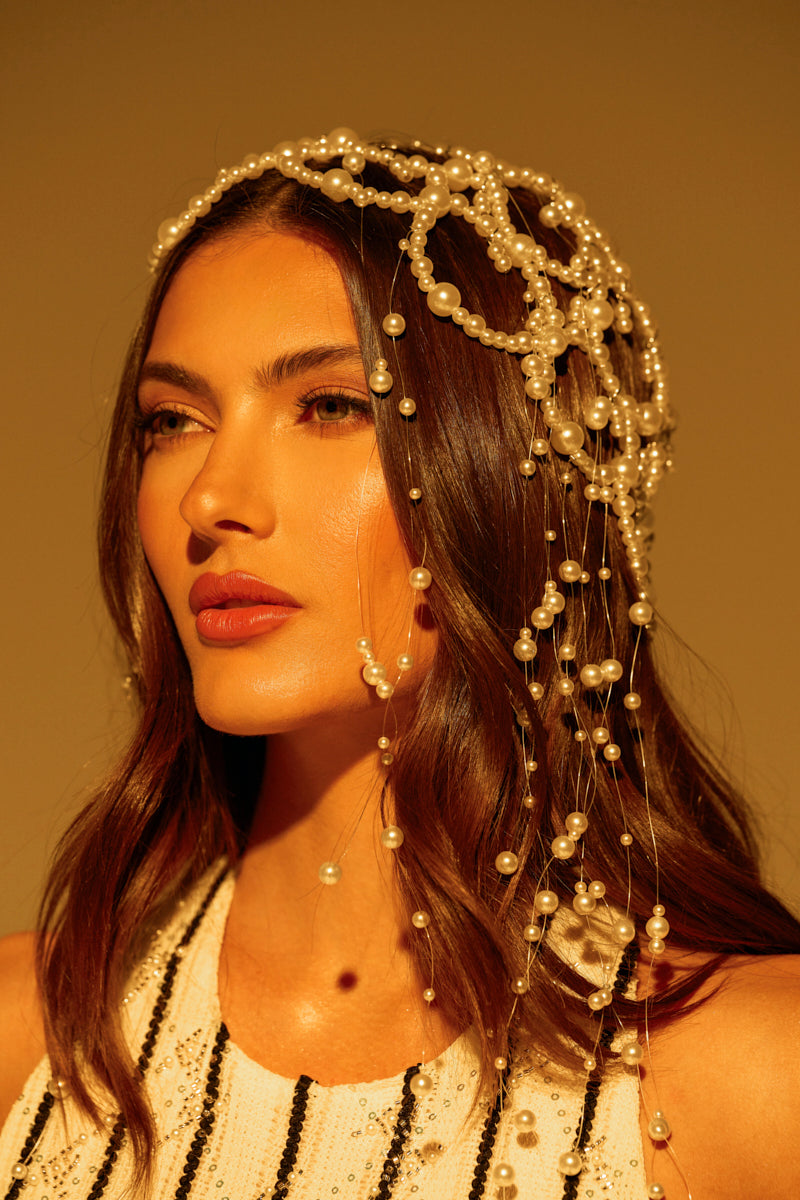 A woman with long, wavy hair wears the elegant PEARL LOVE HEADPIECE, its pearl beads draping down her shoulders. She is in a sleeveless black and white striped outfit, gazing thoughtfully against a warm golden backdrop.