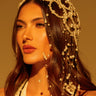 A woman with long, wavy hair wears the elegant PEARL LOVE HEADPIECE, its pearl beads draping down her shoulders. She is in a sleeveless black and white striped outfit, gazing thoughtfully against a warm golden backdrop.