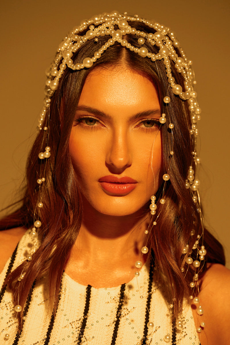 A woman with long brown hair dons the PEARL LOVE HEADPIECE, its strands of pearl beads elegantly draping her face. She wears a white beaded garment featuring a black stripe, set against a warm-toned background that casts a golden glow.